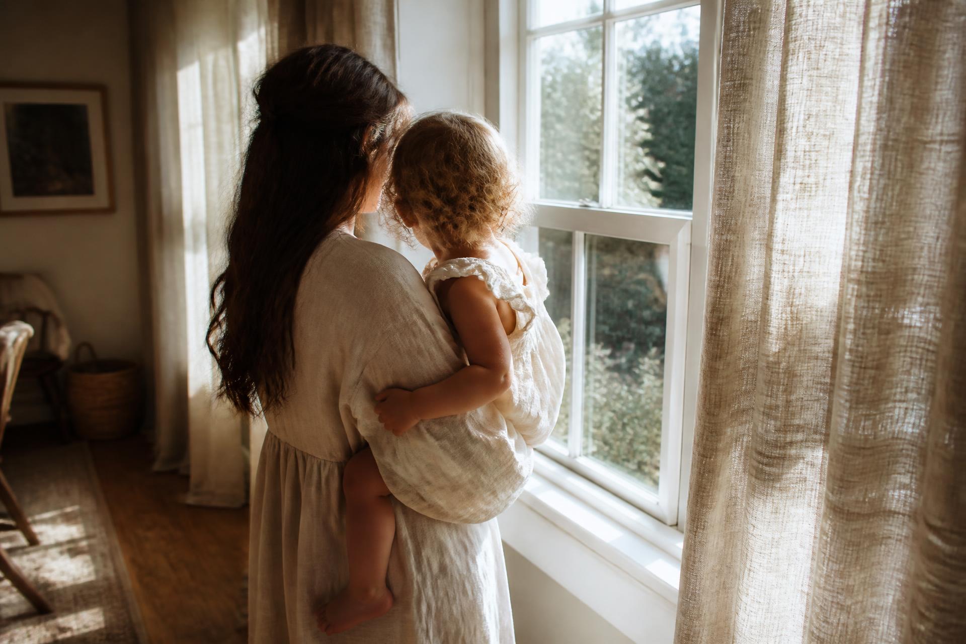 A mother holding her young child by a sunlit window